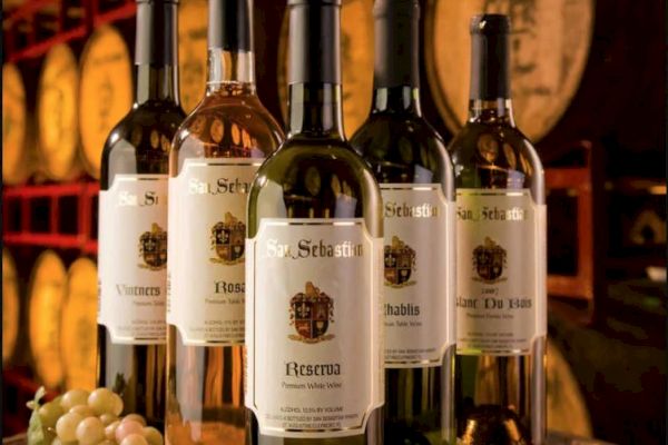Five wine bottles labeled "San Sebastian" on a barrel, surrounded by grapes and corks, with barrels in the background.