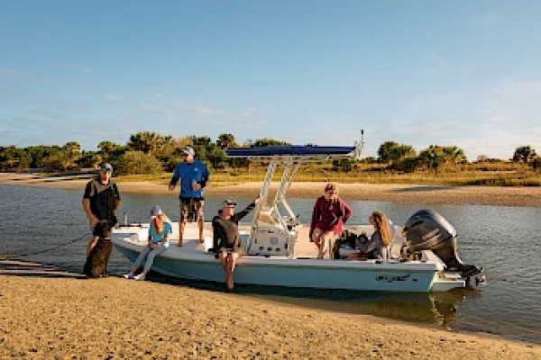 A group of people are on a small boat docked on a sandy shore, enjoying a sunny day by the water with trees in the background.