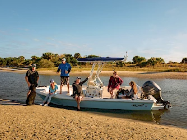 A group of people are on a small boat docked on a sandy shore, enjoying a sunny day by the water with trees in the background.