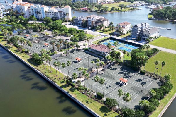 Aerial view of a sports complex with multiple tennis courts, surrounded by greenery and adjacent to a waterway with nearby buildings.