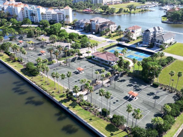 Aerial view of a sports complex with multiple tennis courts, surrounded by greenery and adjacent to a waterway with nearby buildings.