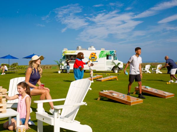 People are enjoying outdoor games and activities on a lawn near a food truck, with chairs, tables, and blue umbrellas scattered around.