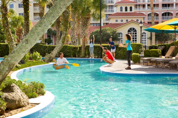 People enjoying a sunny day by a pool. One person is in a cardboard boat while others cheer and watch from the poolside.