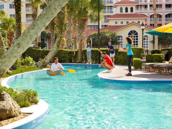 People enjoying a sunny day by a pool. One person is in a cardboard boat while others cheer and watch from the poolside.