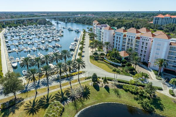 Aerial view of a marina with boats docked, surrounded by palm trees, roads, and a large building with red roofs on the right side.