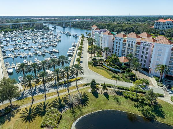 Aerial view of a marina with boats docked, surrounded by palm trees, roads, and a large building with red roofs on the right side.