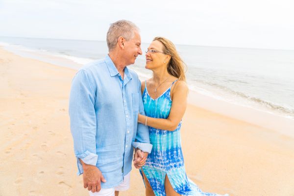 A couple stands on a beach, holding hands and smiling at each other, with the ocean in the background.