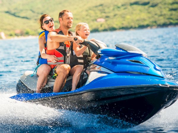 A family of three wearing life jackets rides a blue jet ski on a sunny day, enjoying the water and the scenery.