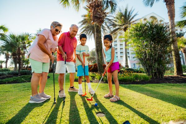 A family of four is playing mini-golf on a sunny day, surrounded by palm trees and a building in the background.