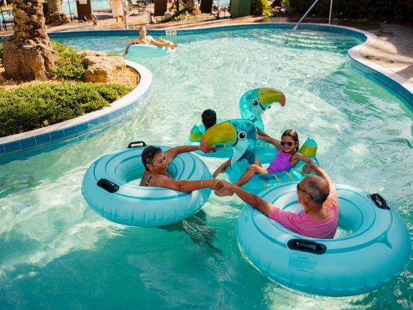 People relaxing on inflatable rings and a parrot-shaped float in a lazy river, surrounded by clear water and lush greenery.