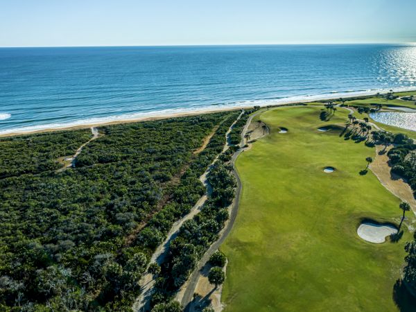 An aerial view of a golf course by the ocean, showing green fairways, sand traps, and trees alongside a beach.