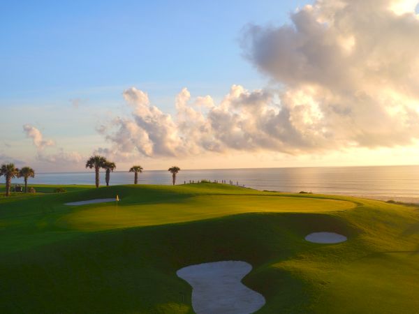 A scenic golf course with palm trees, sand traps, and a view of the ocean under a sky filled with clouds at sunrise or sunset.