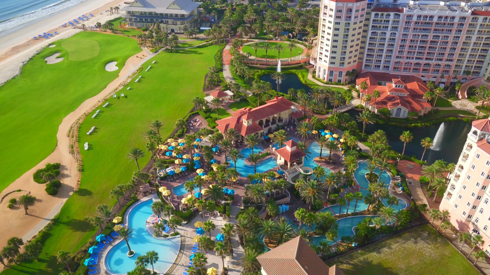 The image shows a resort with pools, palm trees, and nearby beach access, featuring multiple buildings and a golf course.
