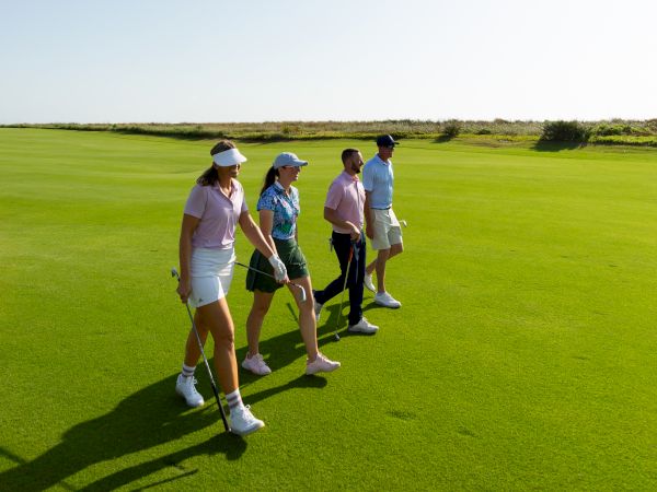 A group of four people walking on a golf course under a clear blue sky, carrying golf clubs and wearing casual sportswear.