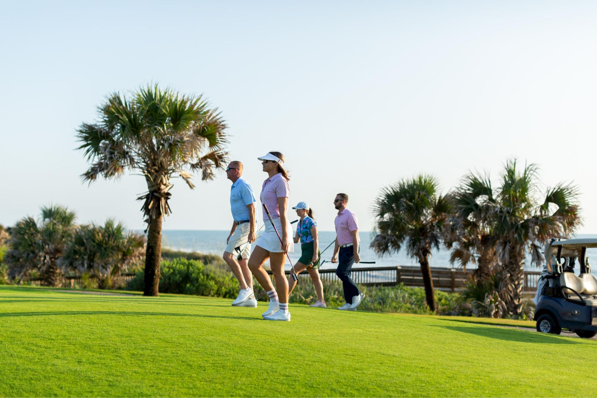 People walking on a golf course with palm trees, near the ocean, and some facing a golf cart under a clear sky.