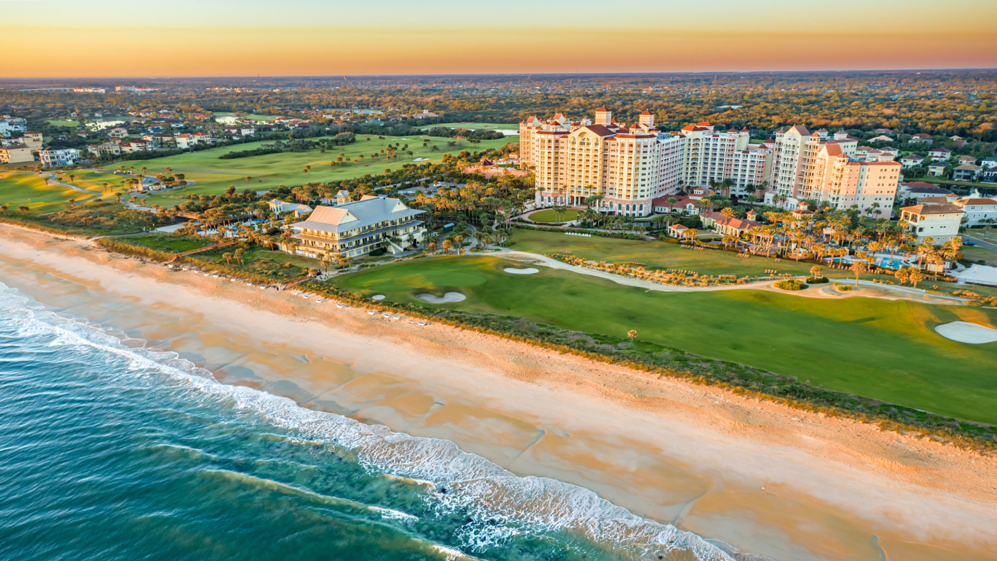 Coastal resort with a golf course, beachfront, and ocean waves; residential area in the background during sunrise or sunset.