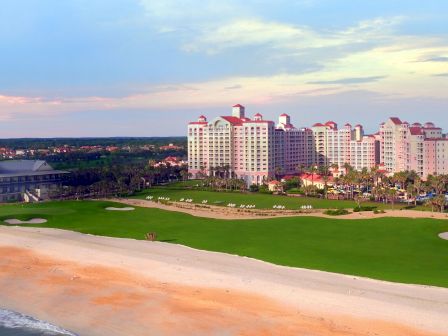 A beachside resort with a golf course, surrounded by palm trees and modern buildings under a colorful sky during sunset.