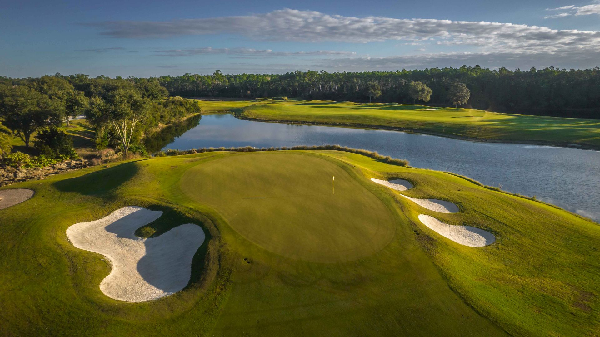 Aerial view of a golf course with green fairways, sand bunkers, and a water hazard surrounded by trees and blue sky.