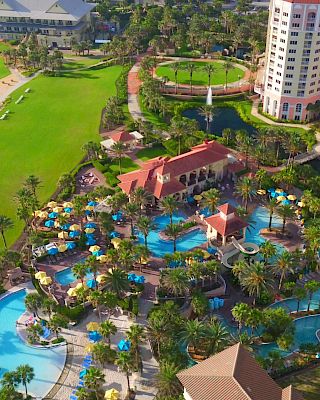 Aerial view of a beachside resort featuring a golf course, multiple pools, sun loungers, and palm trees, with a tall building nearby.
