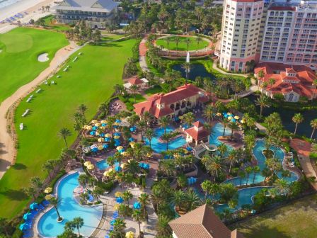 Aerial view of a beachside resort featuring a golf course, multiple pools, sun loungers, and palm trees, with a tall building nearby.