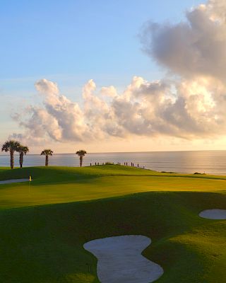 A golf course with palm trees, sand traps, and a flag, overlooking the ocean under a sky with picturesque clouds.