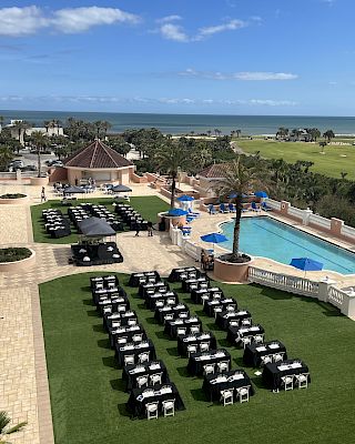 Outdoor setup with rows of tables covered in black cloths on grass, next to a pool. Ocean view in the background with palm trees.