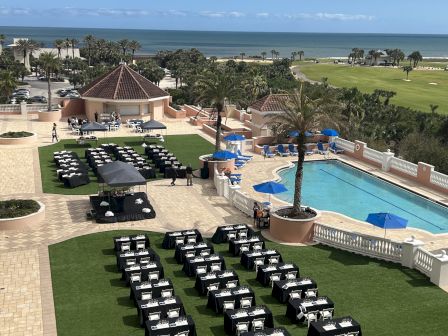 Outdoor setup with rows of tables covered in black cloths on grass, next to a pool. Ocean view in the background with palm trees.
