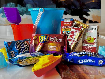 A colorful assortment of snacks and cookies piled together, including Oreos, Doritos, Chips Ahoy, Ditz Bits, Horizon juice, and a yellow toy boat in front, bright cups and a blue scoop behind. ending punctuation.