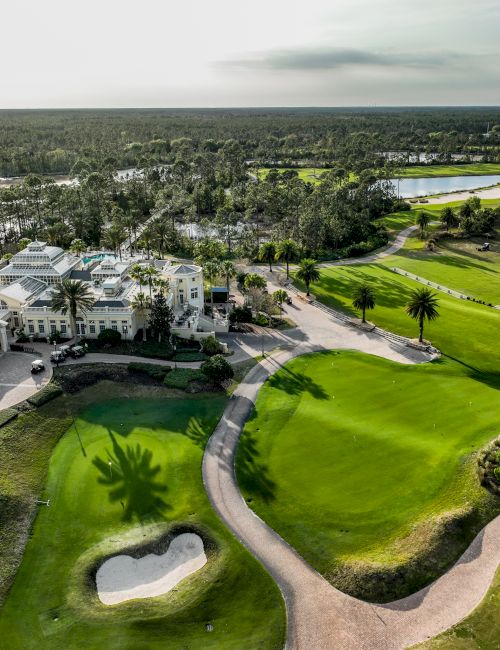 Aerial view of a golf course with a large clubhouse, surrounded by lush greenery, palm trees, and a small water body in the background.