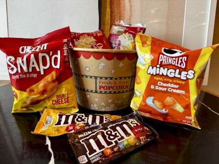 Assorted snacks: Snaps, Doritos-style Nacho Cheddar, Pringles, popcorn cup, and Hi-Chew/Haribo-style bars in front, all on a kitchen counter.