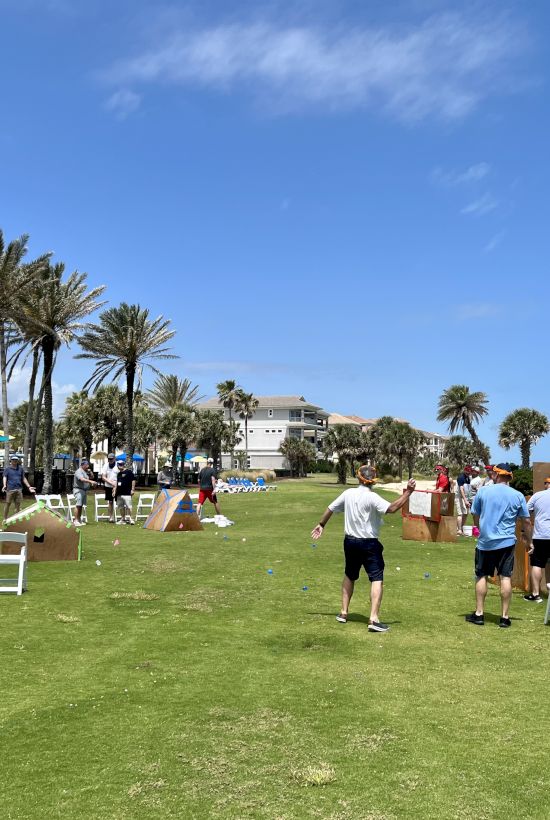 People outside on a grassy area with palm trees, playing a game with chairs and boxes under a clear blue sky.