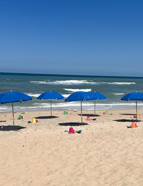 A beach scene with blue umbrellas, colorful beach toys, and gentle waves under a clear blue sky.