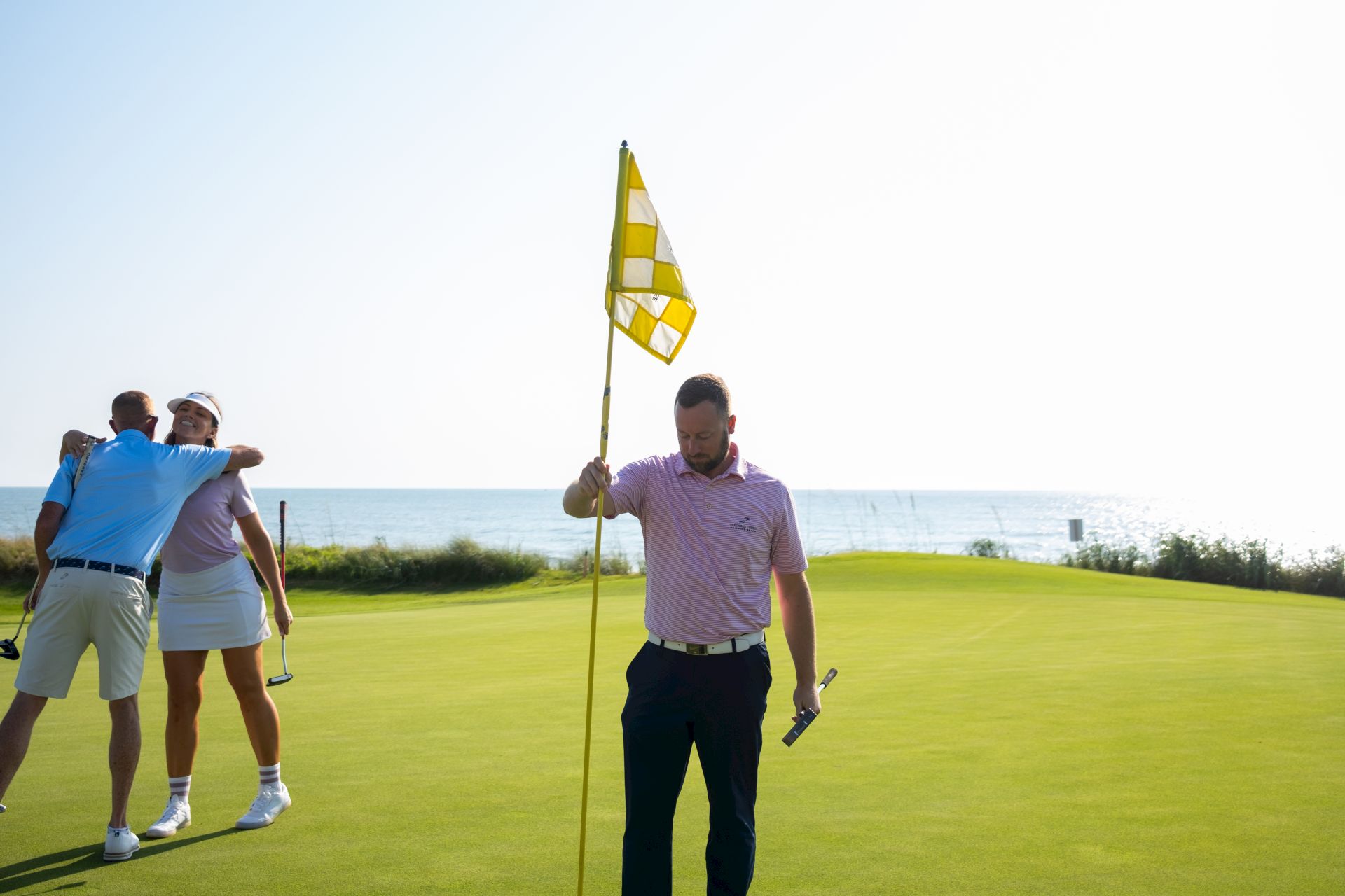 A person holding a golf flag on a course by the sea, with two others embracing in the background.
