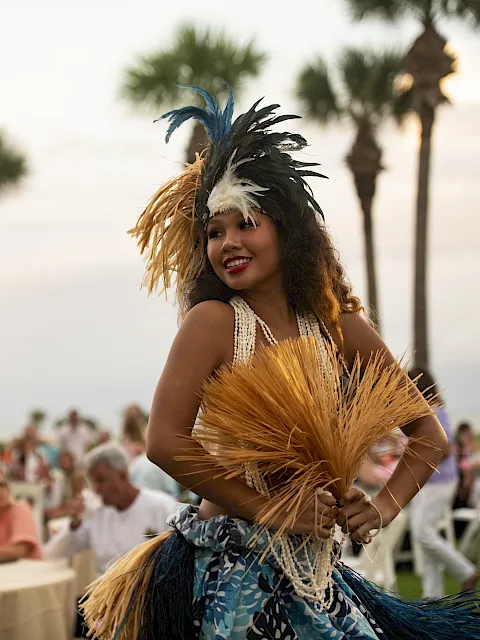 A dancer in a feathered headdress and grass skirt performs at an outdoor event with a seated crowd in the background.