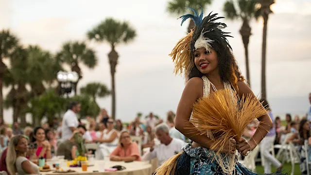 A dancer in a feathered headdress and grass skirt performs at an outdoor event with a seated crowd in the background.