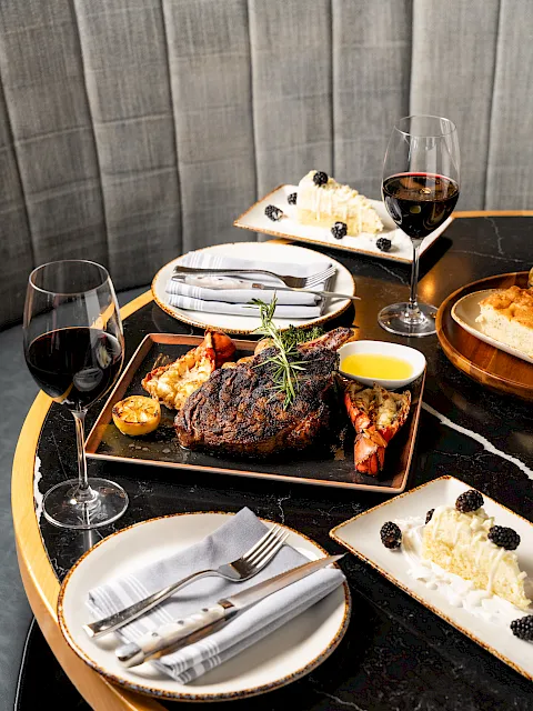 A fine-dining plate with grilled steak, lobster, lemon, herbs, two glasses of red wine, and cheesecake topped with berries.