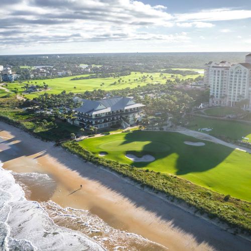 A coastal golf course and luxury resort with a beachfront and ocean waves visible under a partly cloudy sky.