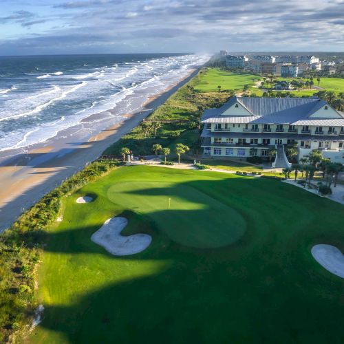 Aerial view of a coastal golf course with sand bunkers, ocean waves, a beach, and a clubhouse surrounded by greenery and palm trees.