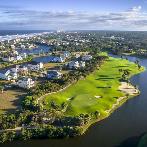 Aerial view of a coastal neighborhood with houses, waterways, and a golf course, all adjacent to the ocean under a partly cloudy sky.