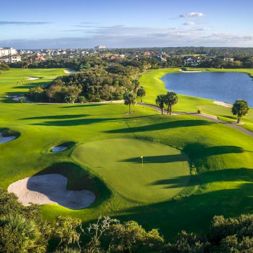 Aerial view of a golf course with sand bunkers, palm trees, a pond, and houses in the distance under a blue sky.