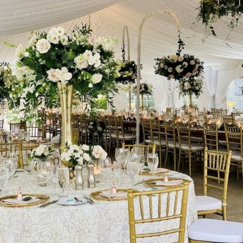 The image shows an elegantly decorated wedding reception with floral centerpieces, gold chairs, and a white canopy tent.