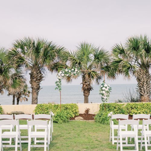 Outdoor beach wedding setup with white chairs, palm trees, and a decorated arch against an ocean backdrop.