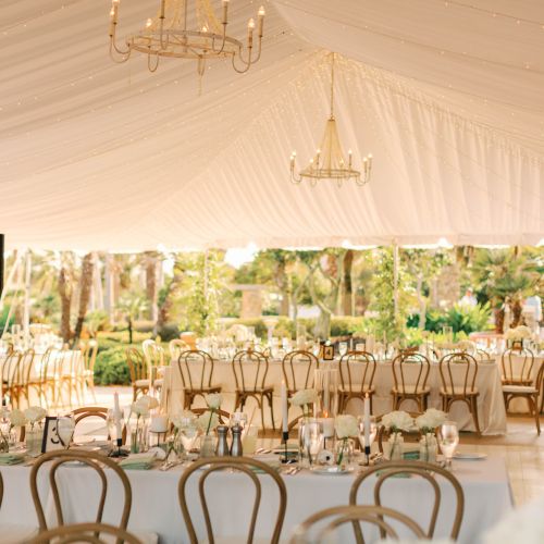Elegant outdoor event setup with draped tent, chandeliers, and decorated tables surrounded by greenery, suggesting a wedding or party.