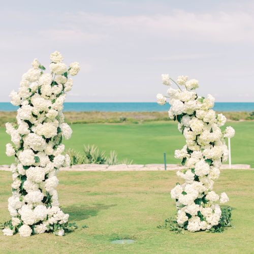 Two tall floral arrangements with white blossoms on a grassy lawn by the sea, under a clear blue sky.