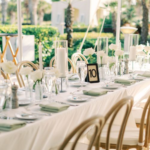 Elegant outdoor dining setup with a long table, white roses, candles, and a "10" table number. Chairs line each side.