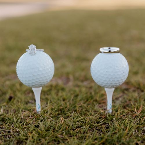 Two golf balls on tees with rings: one ball with a diamond ring, the other with a plain band, placed on grass.