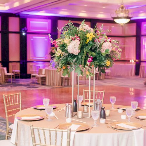 Elegant banquet setting with a round table, tall floral centerpiece, gold chairs, and ambient purple lighting in a spacious room.