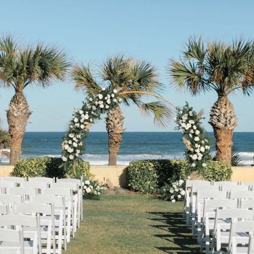 Outdoor wedding setup with white chairs facing an ocean view, featuring a floral arch and palm trees.
