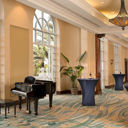 A room with tall windows, a black piano, cocktail tables, a plant, and elegant chandeliers, creating a sophisticated atmosphere.