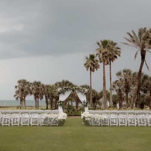 An outdoor wedding setup with white chairs and an arch adorned with flowers, surrounded by palm trees under a cloudy sky.